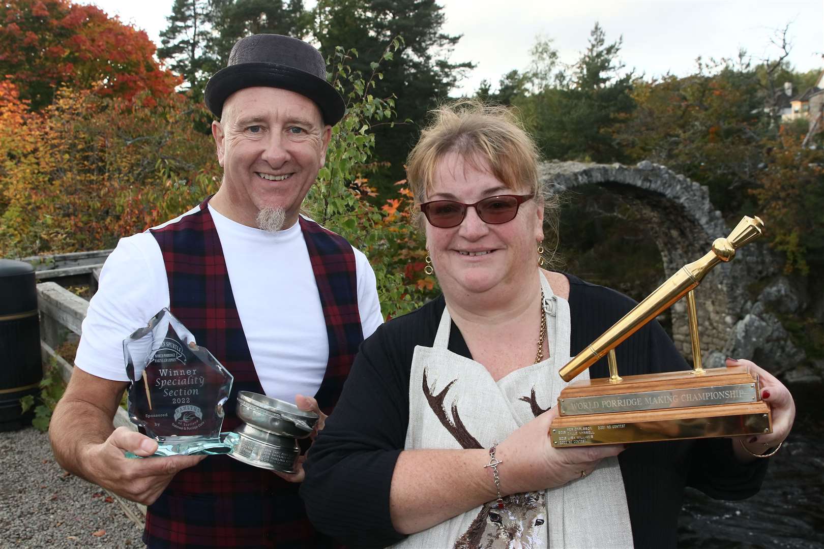 World porridge-making champions for 2022 Chris Young and Lisa Williams pose with their trophies in front of Carrbridge's famous bridge.