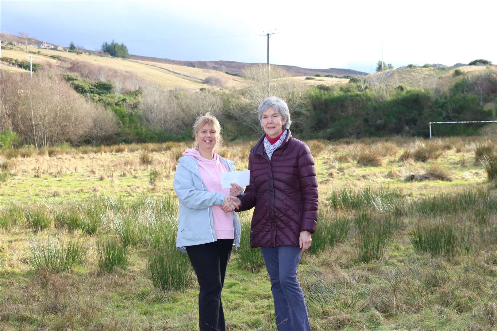 Sarah Fox receives a £1,000 donation towards the Tongue MUGA project from Karen Kelly of Melness Crofters Estate. Behind them is the football pitch where the MUGA will be built.